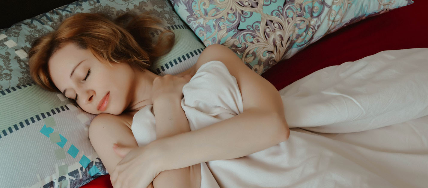 Image shows a young redheaded woman laying happily in bed with the covers pulled up and her eyes closed